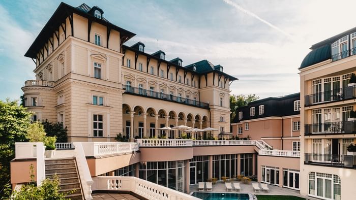 Hotelgebäude mit Pool, Terrasse, Liegen und Stufen, umgeben von Bäumen unter einem blauen Himmel.