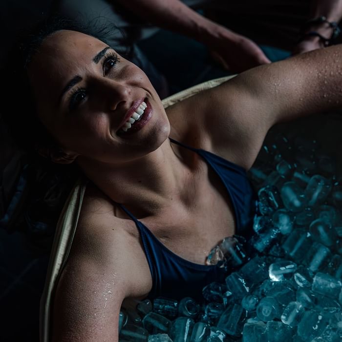 Frau in einer Eiswanne mit einem Lächeln, umgeben von Eiswürfeln und Wassertropfen auf ihrer Haut.