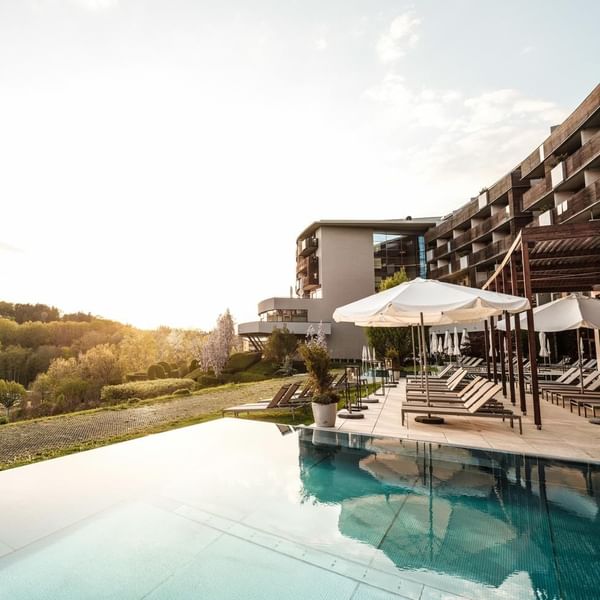 Luxus-Hotel mit Pool, Sonnenliegen, weißen Sonnenschirmen und umgeben von Bäumen bei Sonnenuntergang.