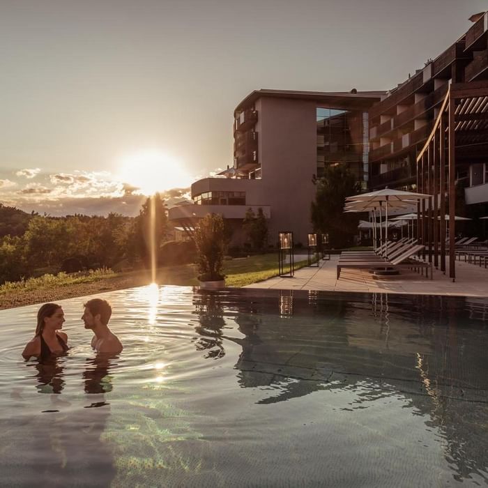 Zwei Personen genießen einen Sonnenuntergang im Pool vor einem modernen Hotelgebäude.