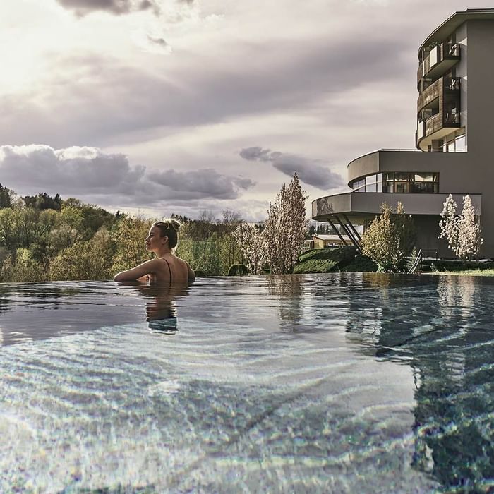 Eine Frau entspannt sich im Schwimmbad vor einem modernen Gebäude mit Bäumen im Hintergrund.